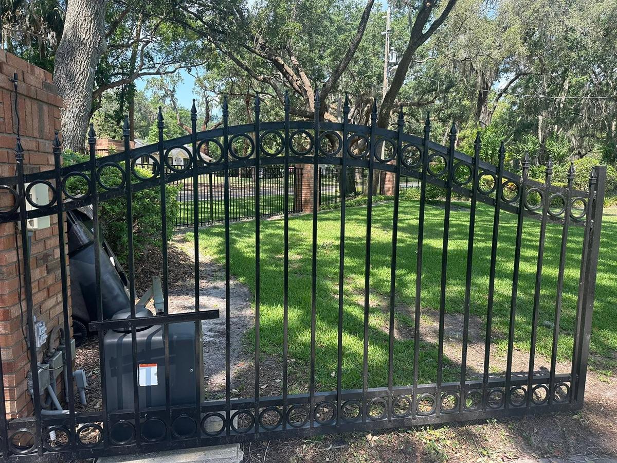 Black arched steel driveway gate with ring detailing across a tree-lined drive