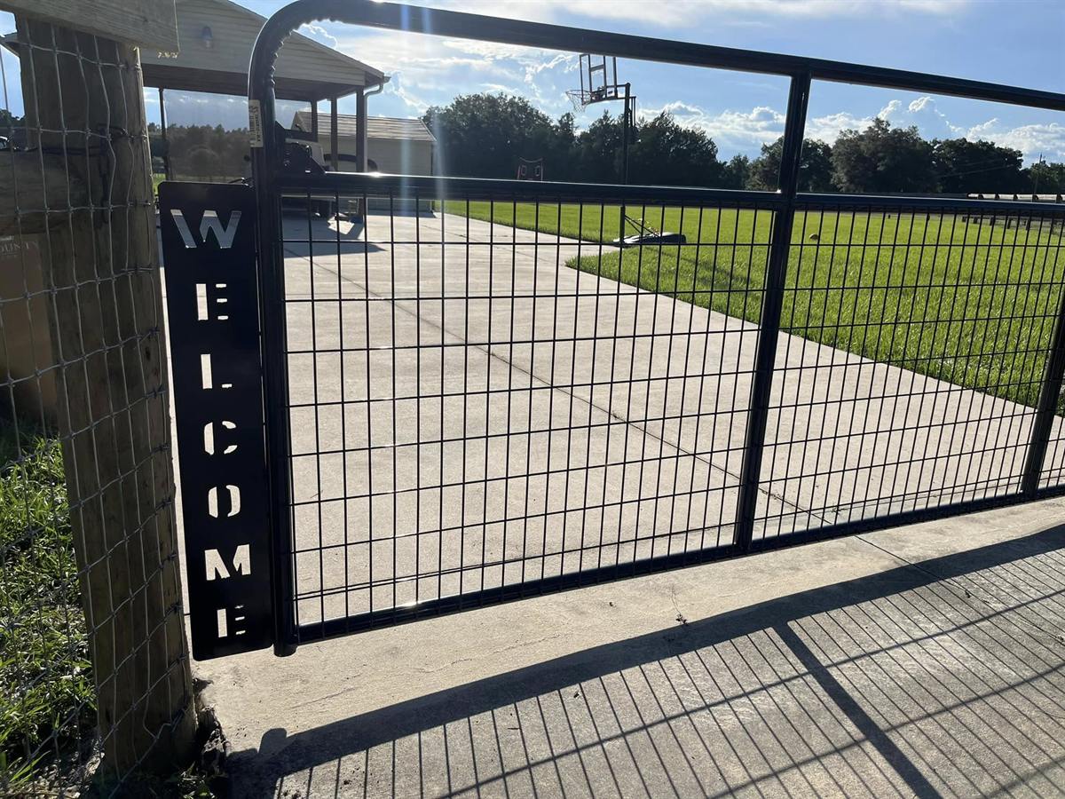 Black steel gate with welded wire panel and a vertical WELCOME sign post at a driveway